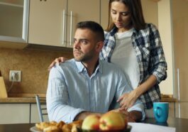 Woman comforts man at kitchen table with food.