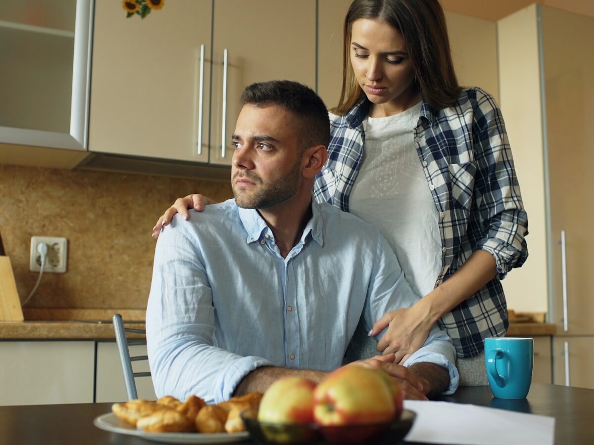 Woman comforts man at kitchen table with food.