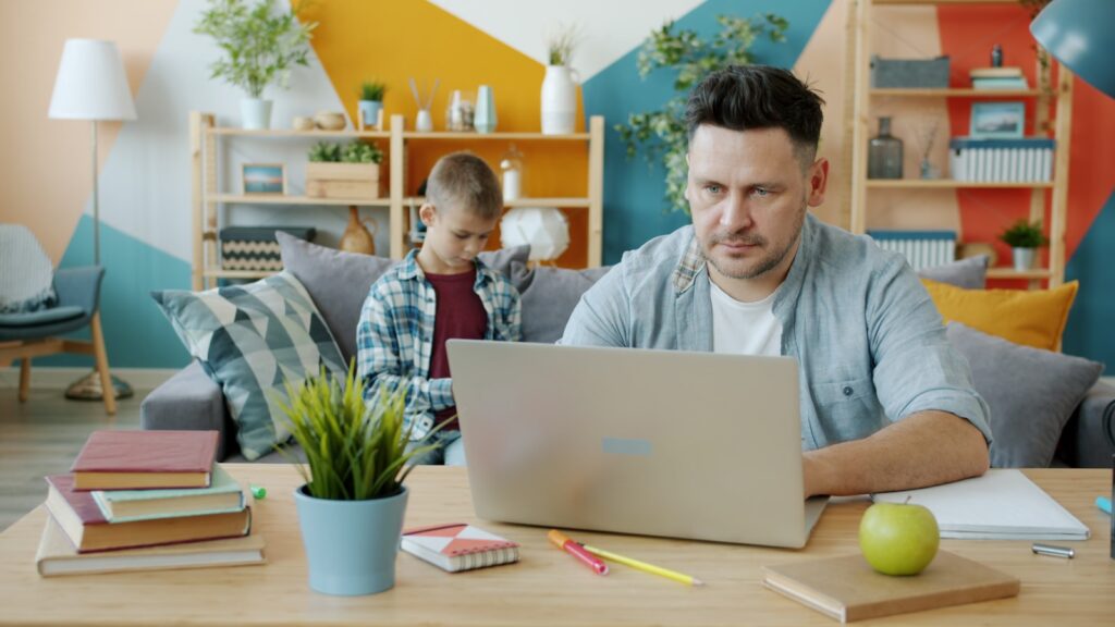 Father working on laptop while son uses phone on couch.
