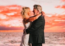 a bride and groom embracing on the beach at sunset