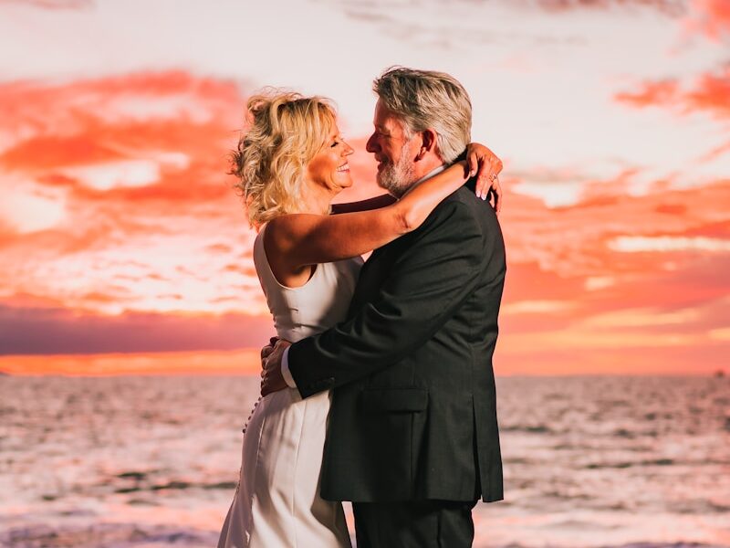 a bride and groom embracing on the beach at sunset
