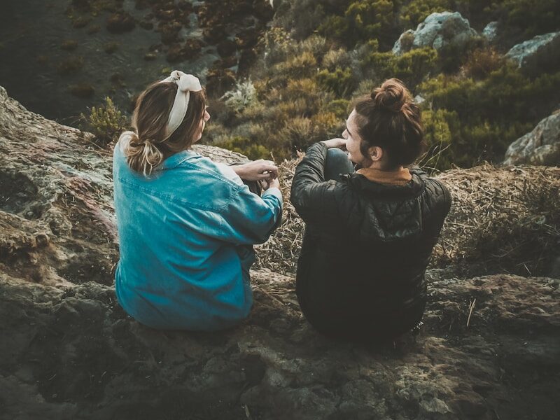 two women sitting on cliff while chatting viewing blue body of water