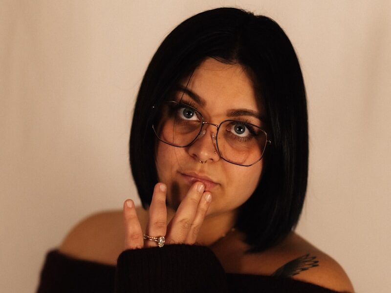 Woman with short dark hair and glasses touching face.