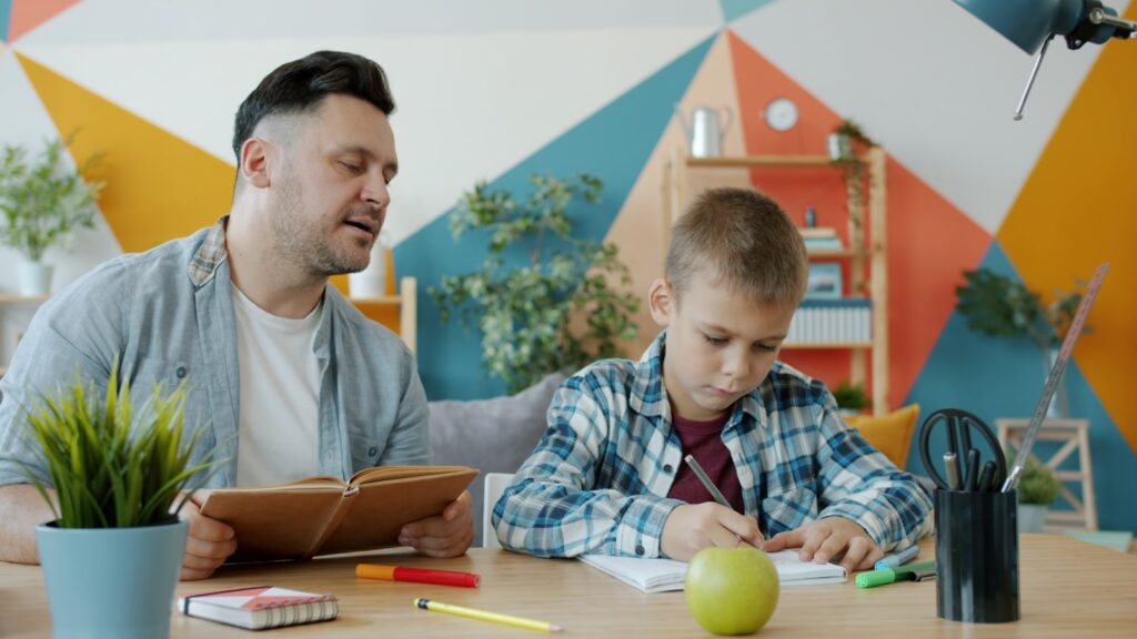 Father helps son with homework at a colorful desk.