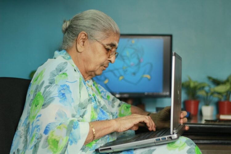 Elderly Indian woman in floral saree using a laptop at home.