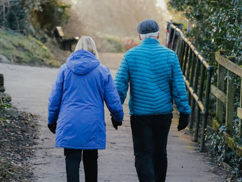 A senior couple in winter jackets walking together in a park pathway at sunset.