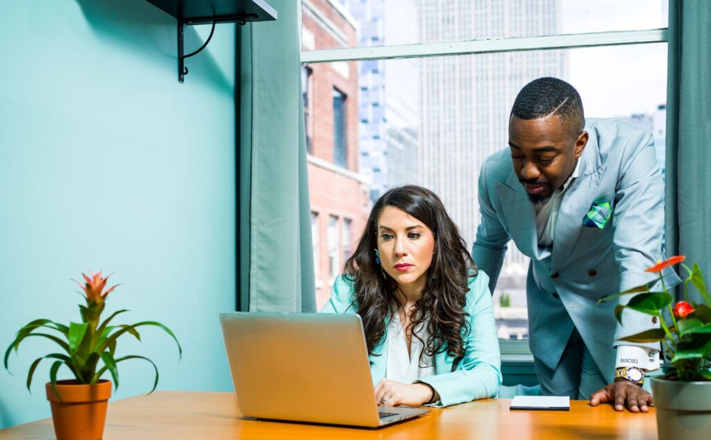 Two colleagues work together in a modern office setting, focusing on a laptop.