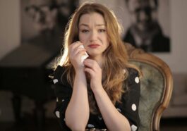 A woman sitting in a vintage chair indoors, showing emotions and holding tissues.