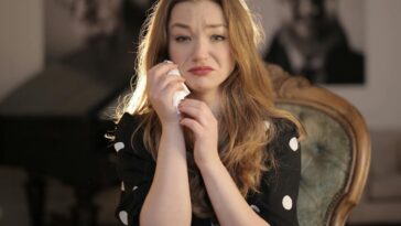 A woman sitting in a vintage chair indoors, showing emotions and holding tissues.