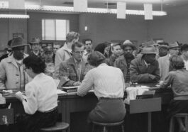 Unemployed men, including African Americans, line up at a counter for assistance from women office workers at an unemployment office, Detroit, Michigan.