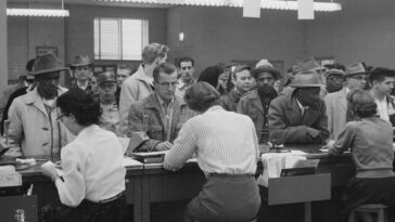 Unemployed men, including African Americans, line up at a counter for assistance from women office workers at an unemployment office, Detroit, Michigan.