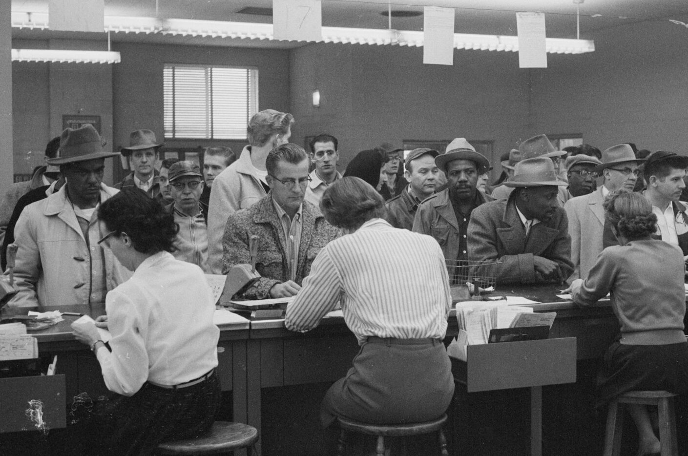 Unemployed men, including African Americans, line up at a counter for assistance from women office workers at an unemployment office, Detroit, Michigan.