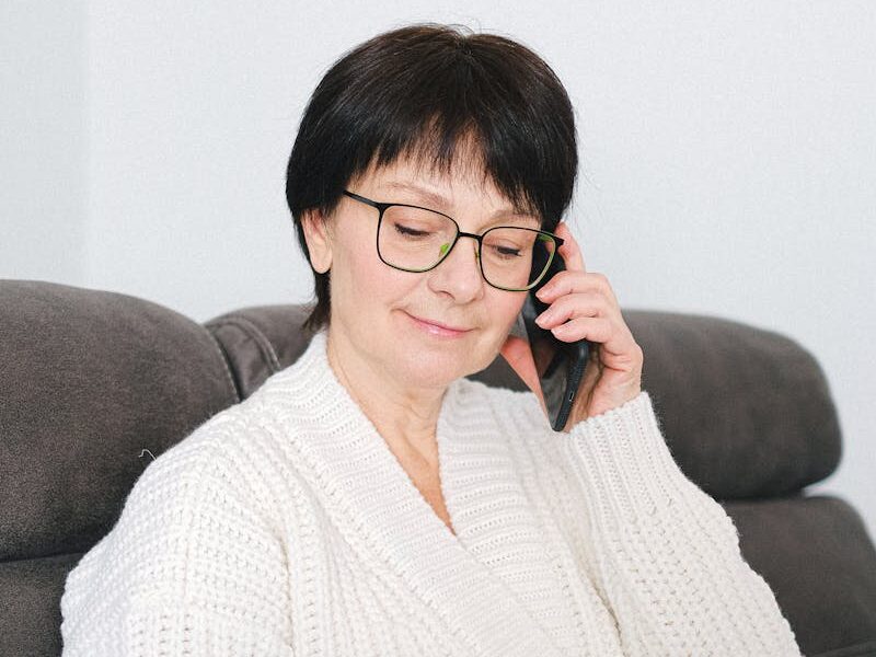 Elderly woman in a cozy sweater using a laptop and phone at home.