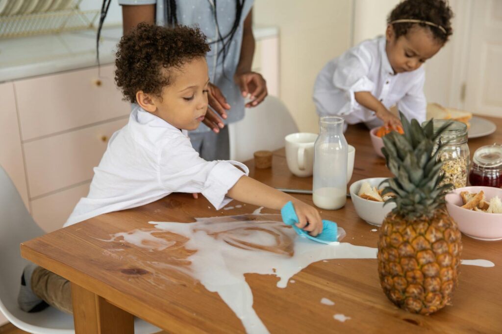 Children clean up a milk spill on a wooden breakfast table with family nearby, creating a cute and lively morning scene.