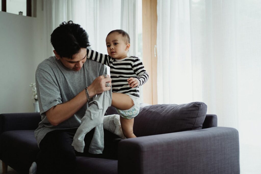 A father lovingly dresses his baby son on a sofa in a bright, cozy living room.