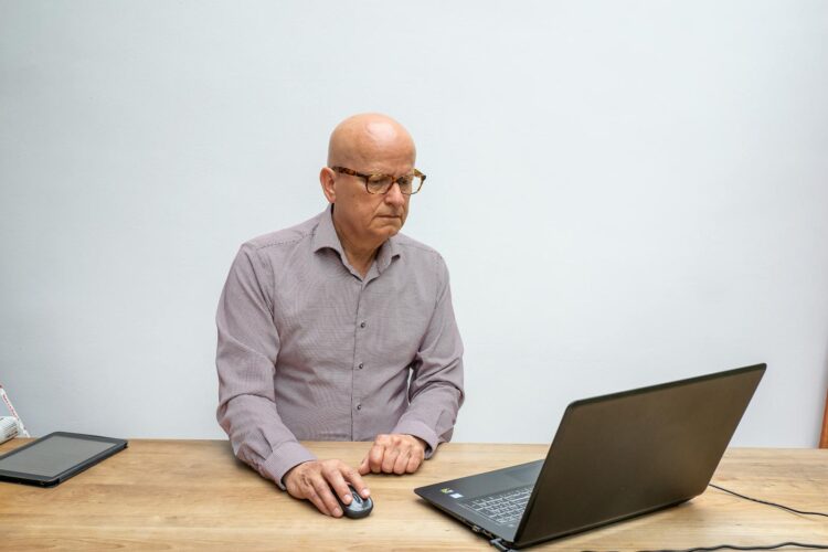 Elderly man using laptop and mouse, focused at work in a bright setting.