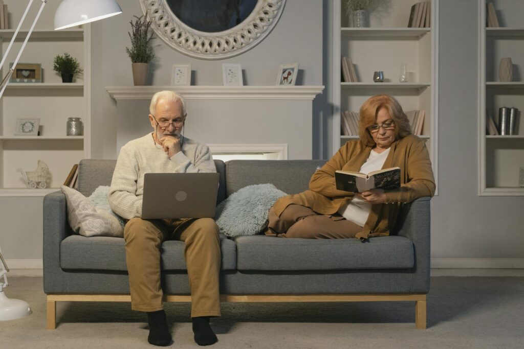 Elderly couple sitting on a sofa at home, man using laptop and woman reading a book, depicting modern senior lifestyle.