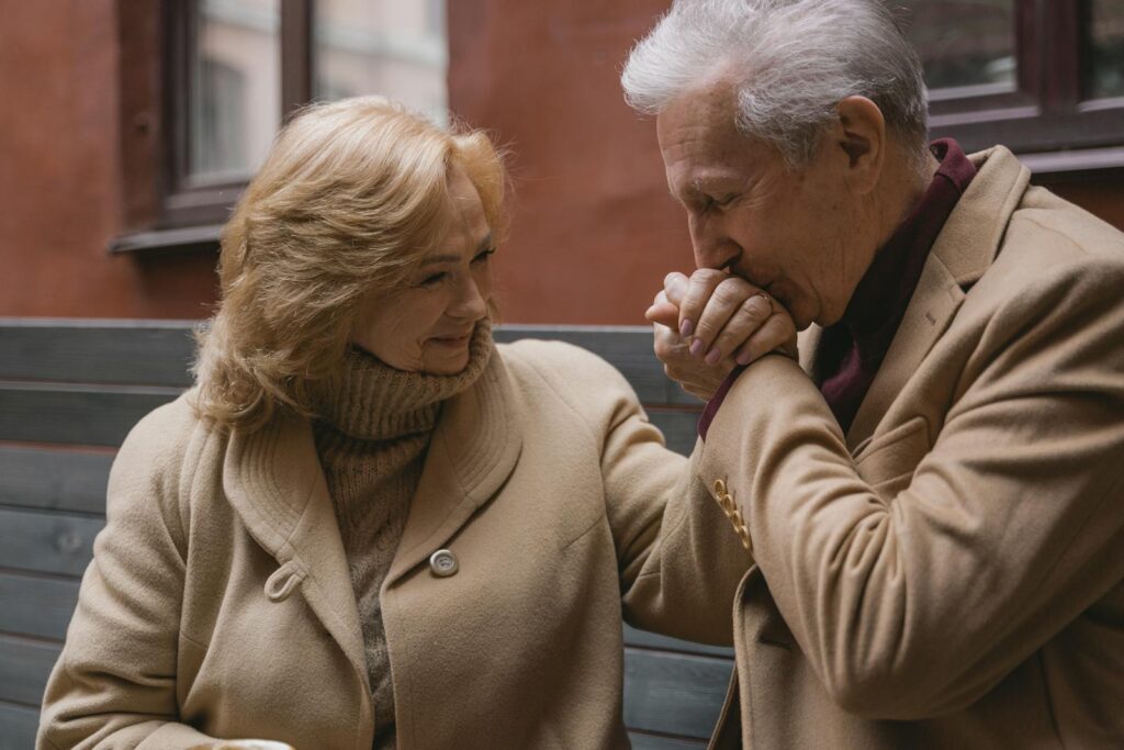 Elderly couple in beige coats sharing a loving moment, holding hands and smiling outdoors.