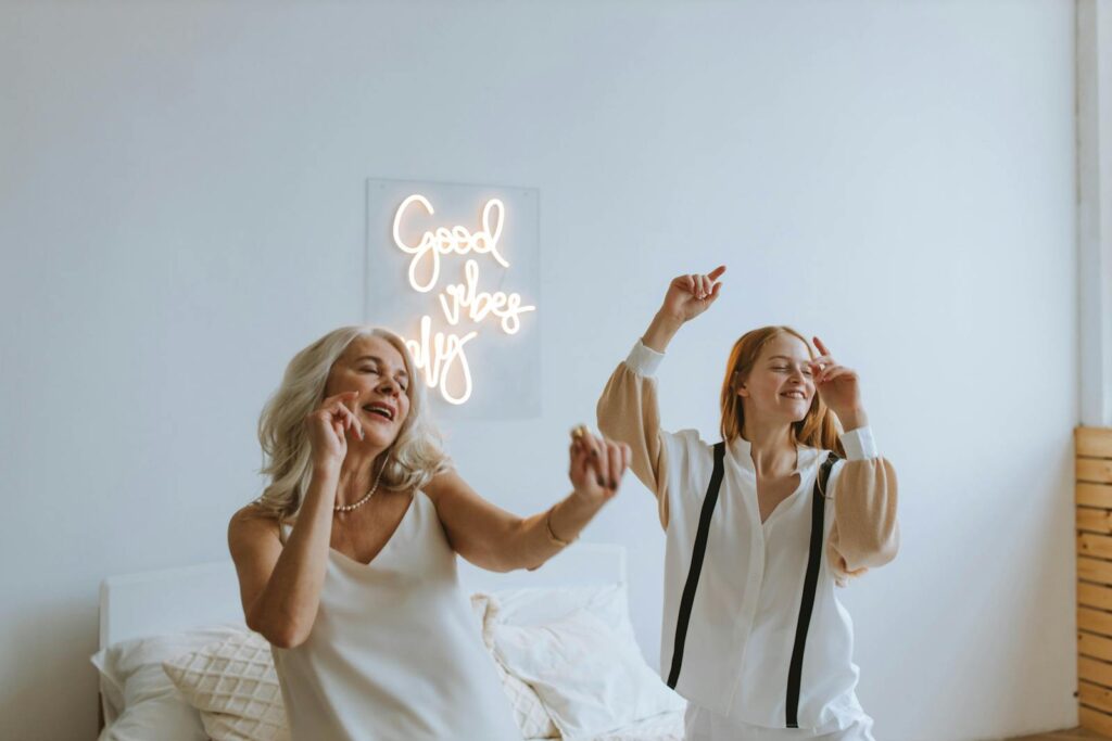 A grandmother and granddaughter dancing joyfully in a cozy room with a 'Good Vibes Only' sign.