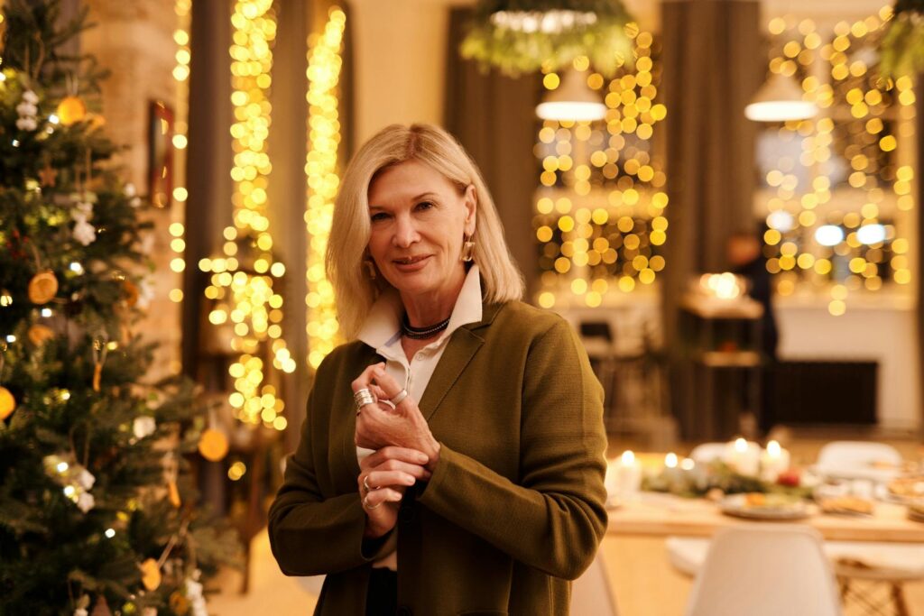 Smiling senior woman in festive setting with bokeh lights and Christmas tree, exuding warmth and joy.