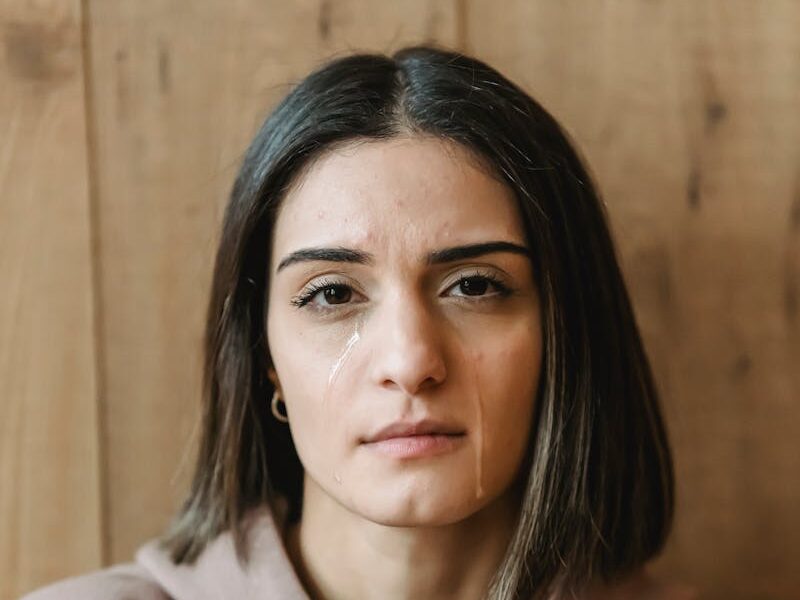Young woman with tears looking serious, sitting indoors against wooden background.