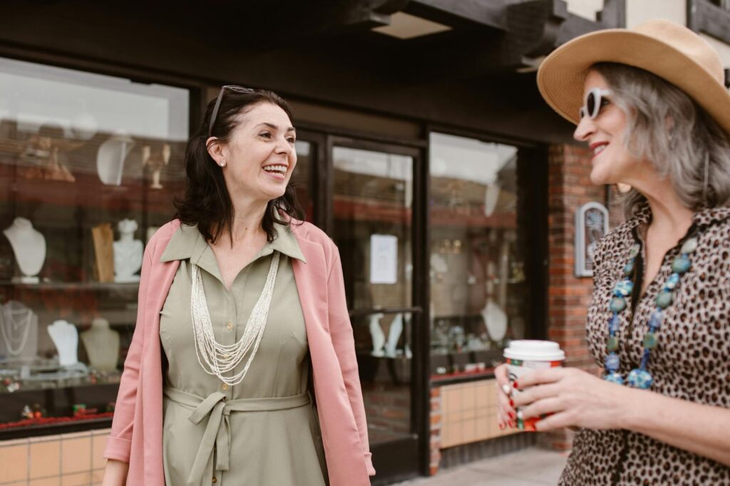 Two Caucasian women smiling and chatting outdoors, one holding a coffee cup.