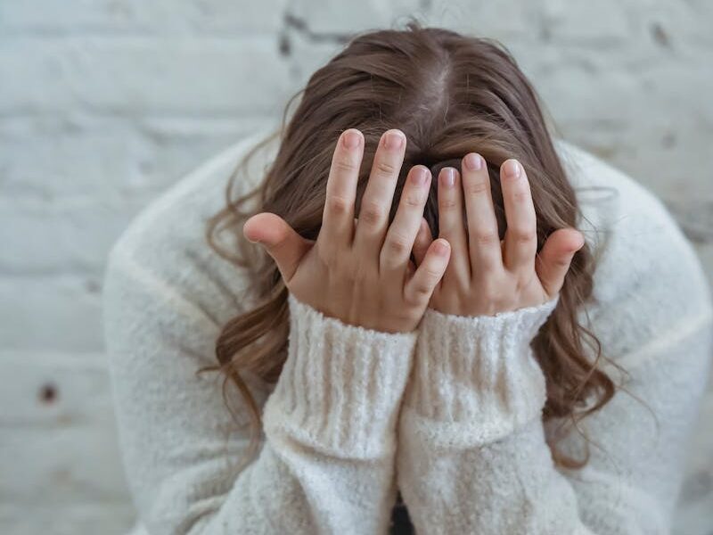 Unrecognizable tired female in casual clothes covering face with hands while sitting on soft surface near brick wall in light apartment