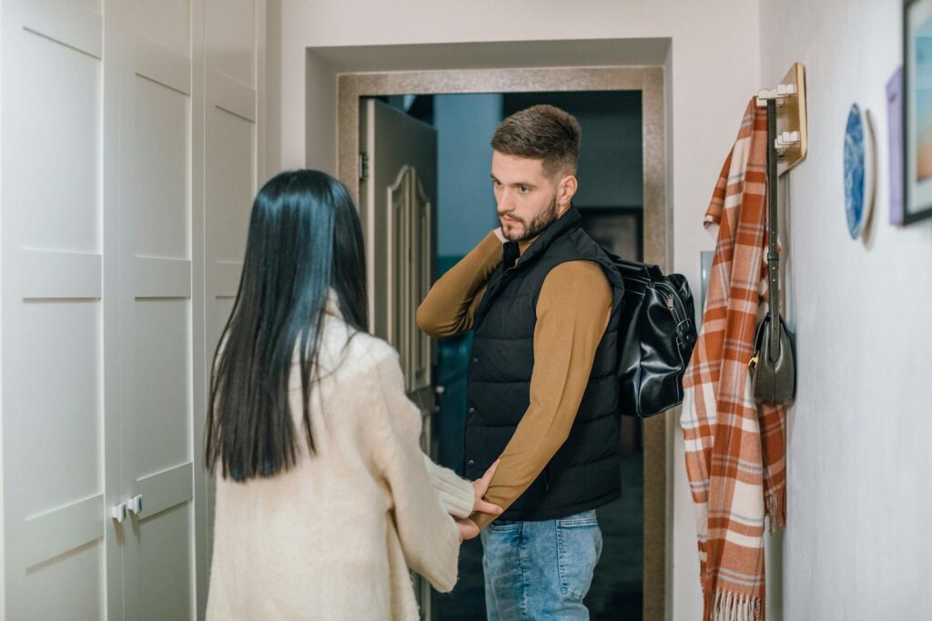 A couple holds hands in a hallway as a man prepares to leave with a backpack.