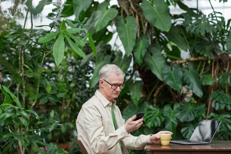 Elderly man engaged in remote work with laptop and smartphone surrounded by lush greenery.