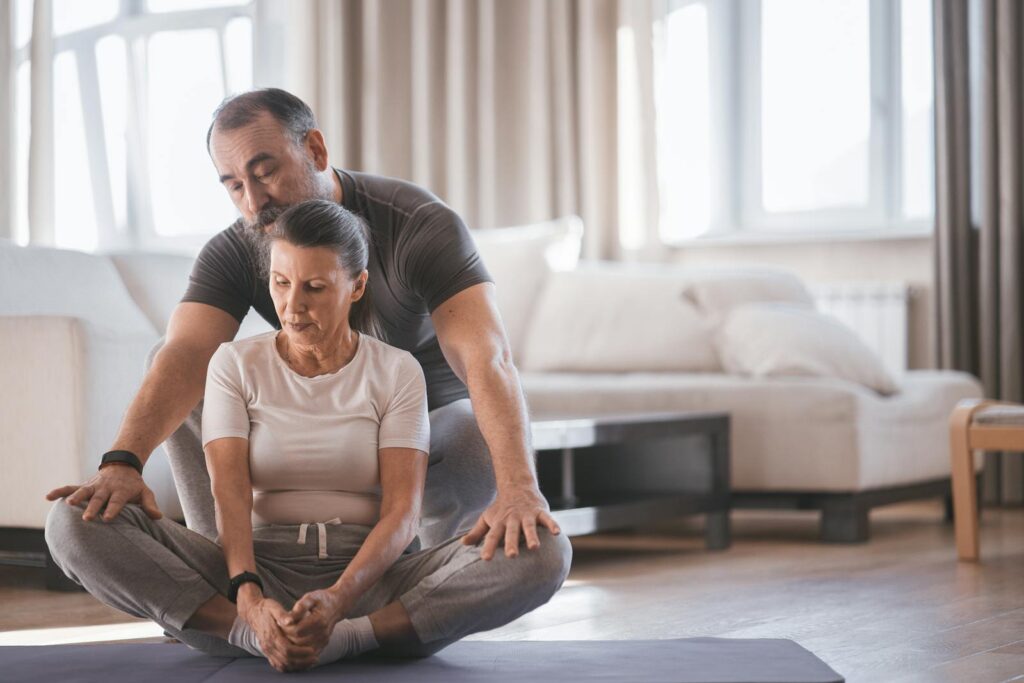 Elderly couple engaging in a yoga session indoors, practicing bound angle pose with focus and support.