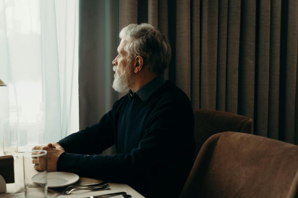 An elderly man with a beard looking outside while sitting by a table indoors.