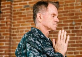 A US Navy soldier in uniform praying with hands clasped against a brick wall backdrop.