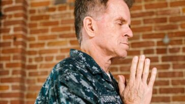 A US Navy soldier in uniform praying with hands clasped against a brick wall backdrop.