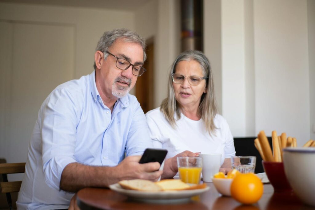 An elderly couple enjoys breakfast together, using a smartphone at a cozy table indoors.