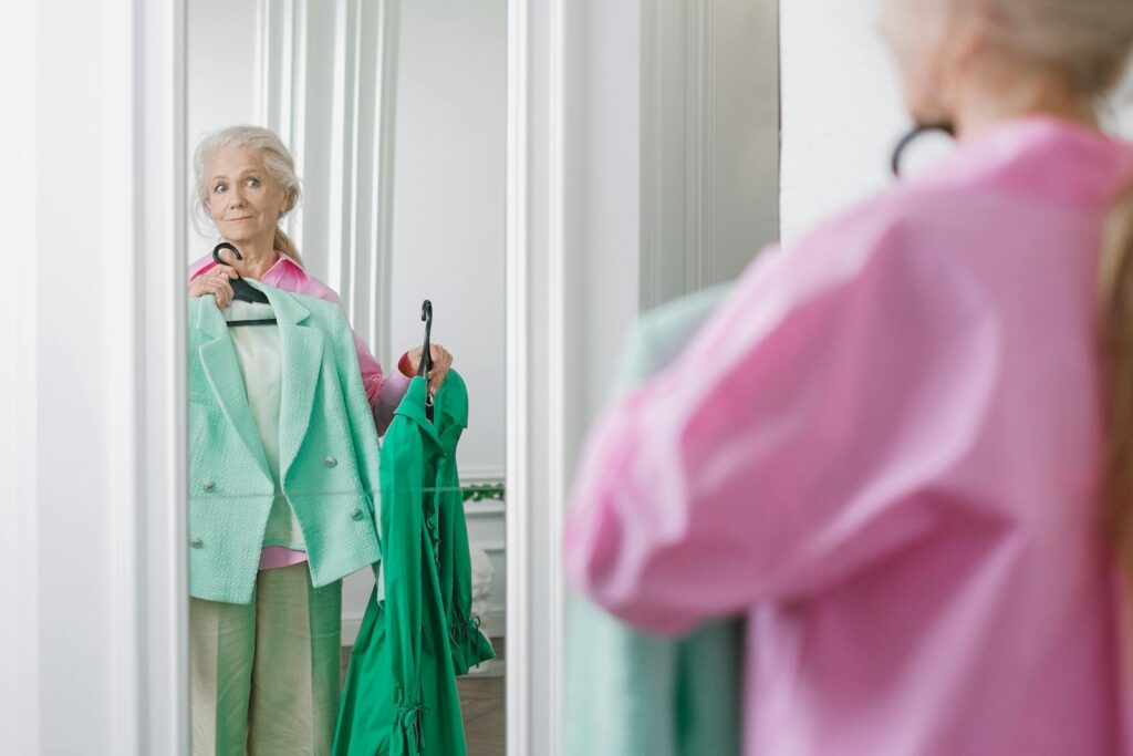 Elderly woman holding clothes in front of mirror, deciding on outfit.