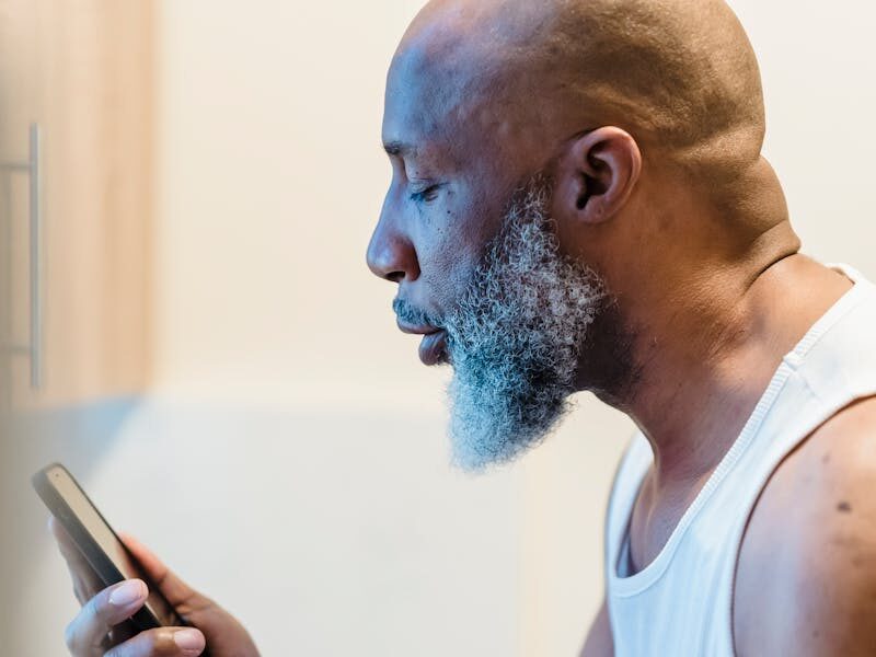 Senior man in kitchen, expressing frustration during a phone call, wearing a white tank top.