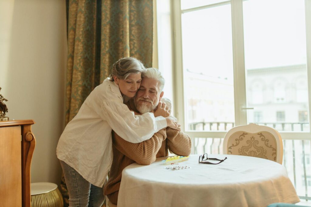A loving elderly couple shares a warm embrace at a dining table indoors, symbolizing affection and togetherness.