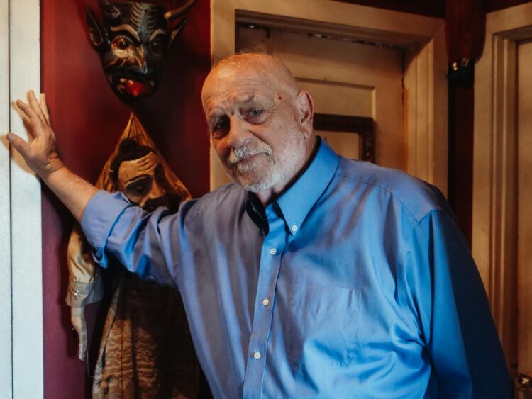 Senior man in a blue shirt leaning against a wall with artistic masks indoors.