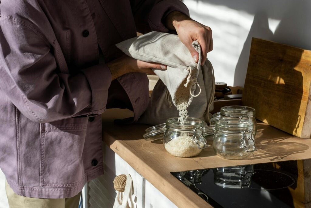 A person pours rice from a cloth bag into a glass jar on a kitchen counter, in natural sunlight.