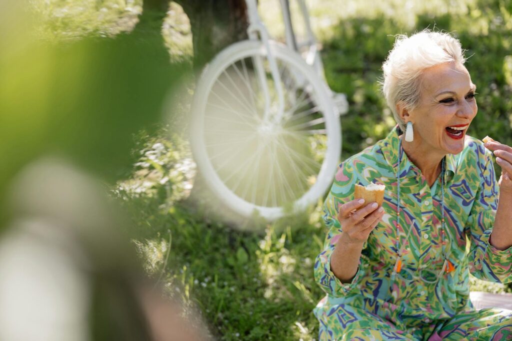 A joyful senior woman eating bread while sitting in a sunny park, next to a bicycle.