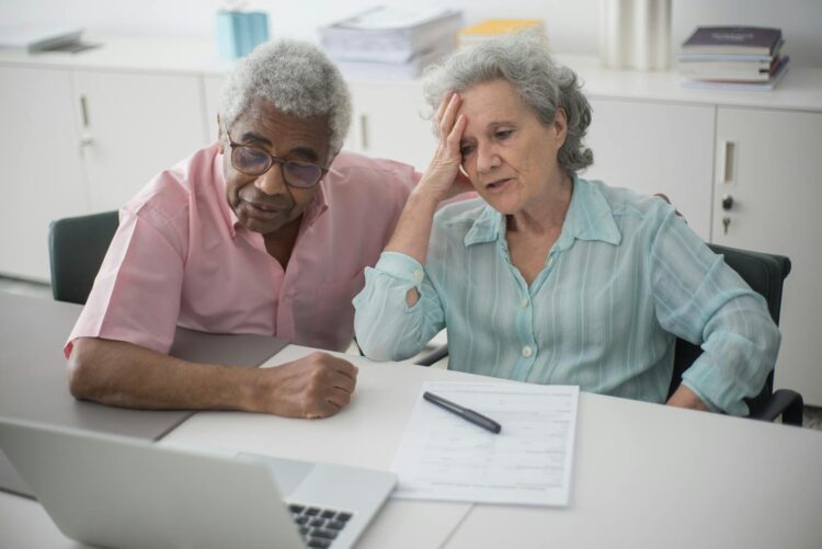 Elderly couple engaged with a laptop, reviewing paperwork at home office desk.