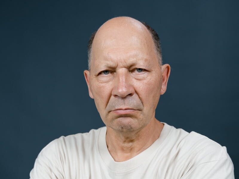 Portrait of a bald elderly man with arms crossed showing intense expression in a studio setting.
