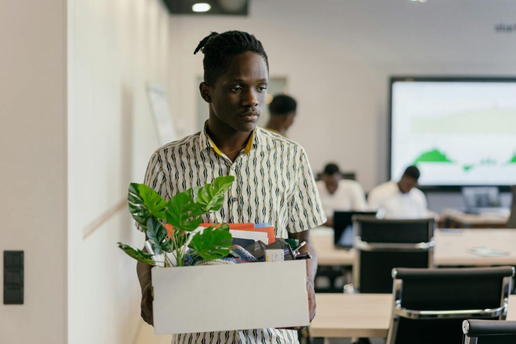 A serious young man carrying a box of personal items in an office setting, suggesting dismissal or resignation.