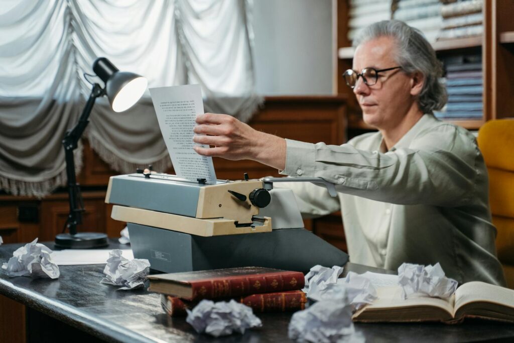 Elderly man with glasses using a vintage typewriter, surrounded by books and papers in a study.