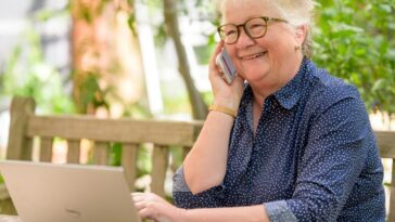 a woman on her phone while sitting at a table with a laptop