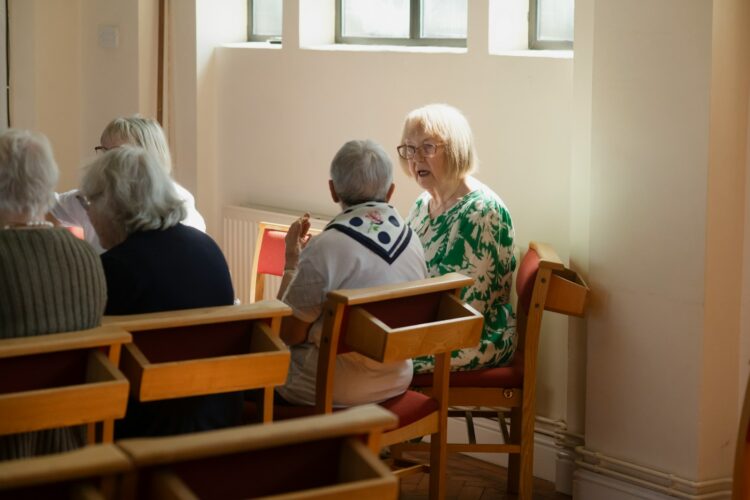 Elderly women sit and chat together indoors.