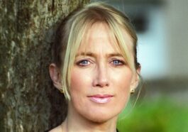 Blonde woman with blue eyes leaning against tree