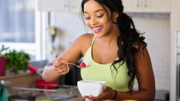 Smiling woman eating a nutritious fruit bowl in a bright kitchen setting.