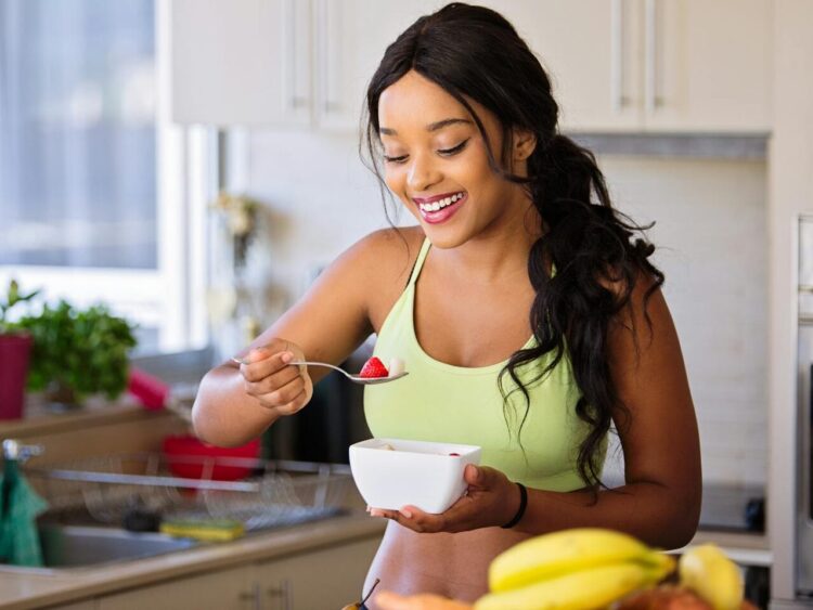 Smiling woman eating a nutritious fruit bowl in a bright kitchen setting.