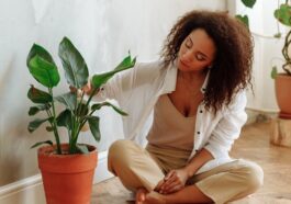 An African American woman sitting on a wooden floor, caring for a potted indoor plant in a bright, modern home.